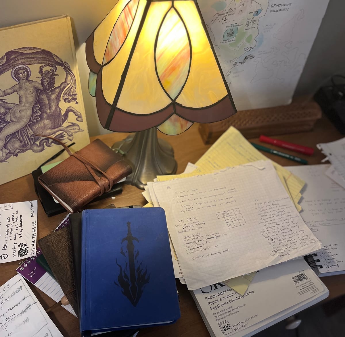 A cluttered desk covered in journals, paper, markets, a stain glass lamp, a cream colored book depicting roman gods